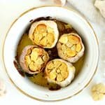 Four heads of roasted garlic with golden, caramelized cloves in a white baking dish surrounded by fresh garlic.