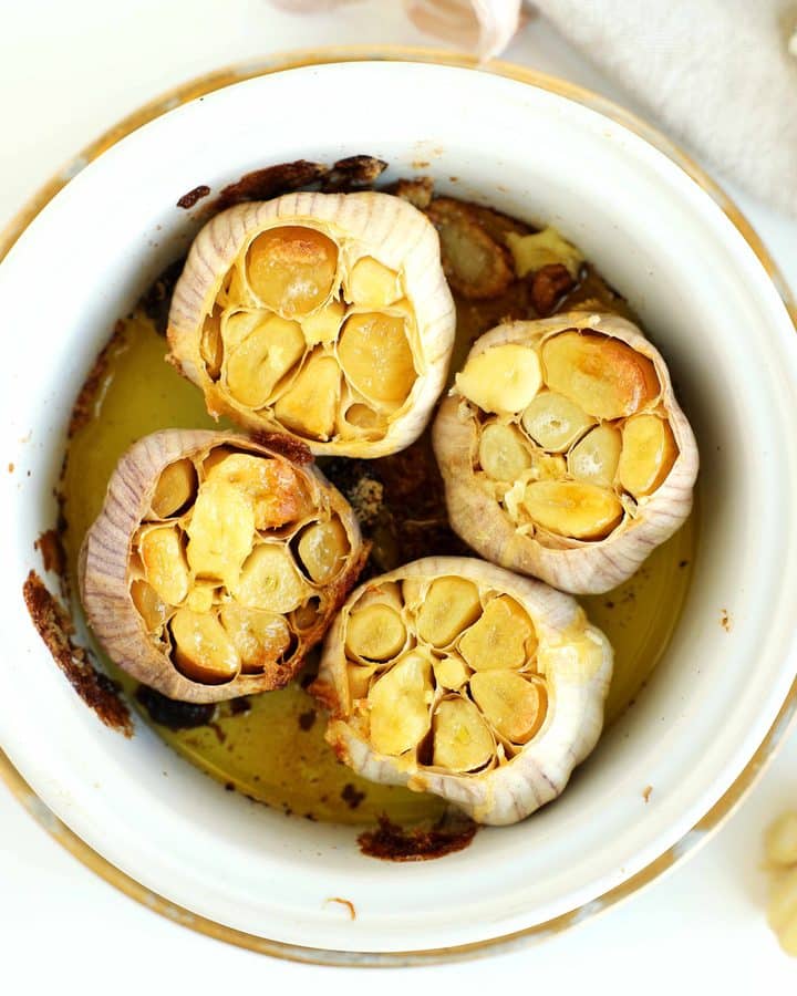 Four heads of roasted garlic with golden, caramelized cloves in a white baking dish surrounded by fresh garlic.