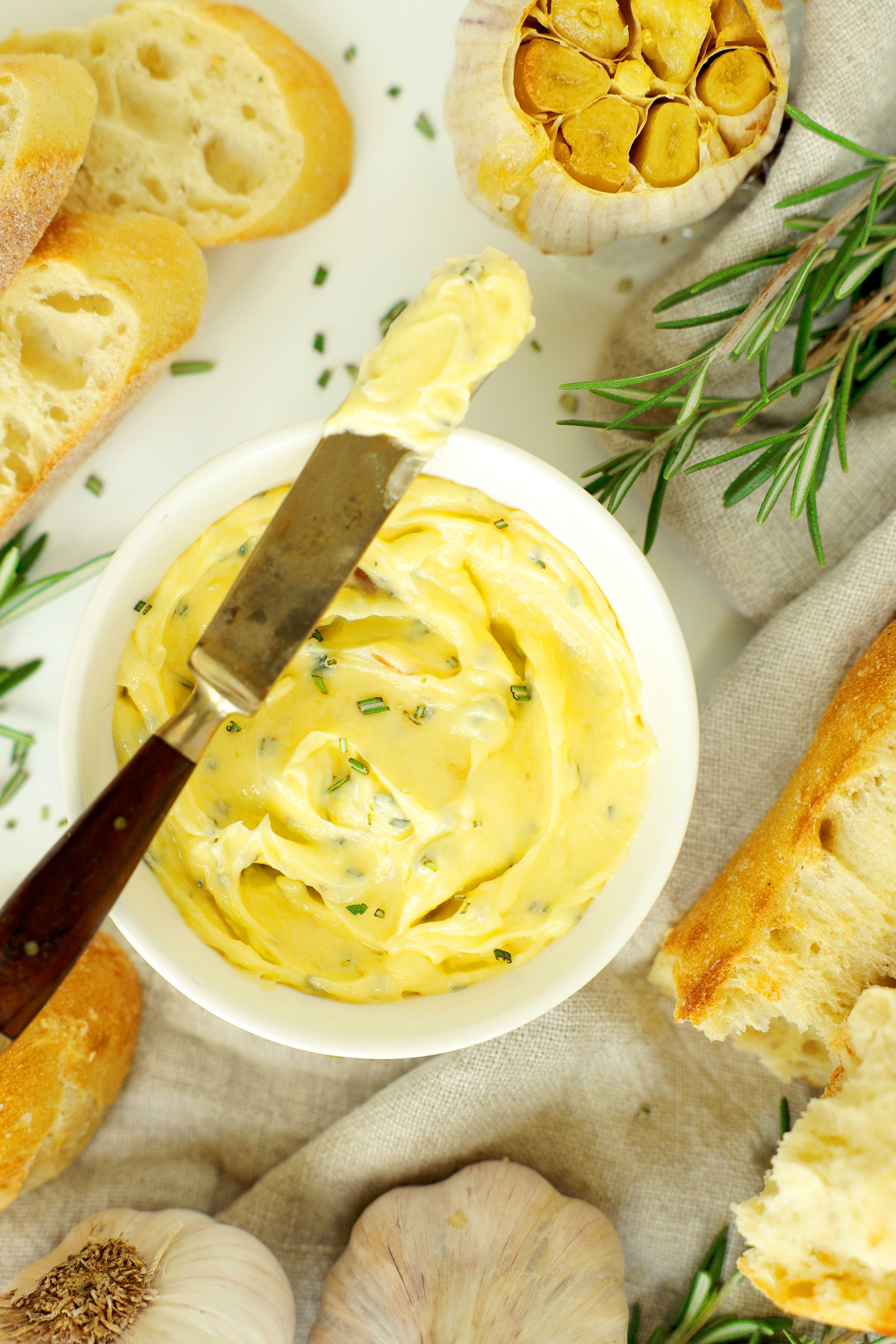 Roasted garlic butter in a white bowl with a butter-covered knife, sliced bread, and fresh rosemary.