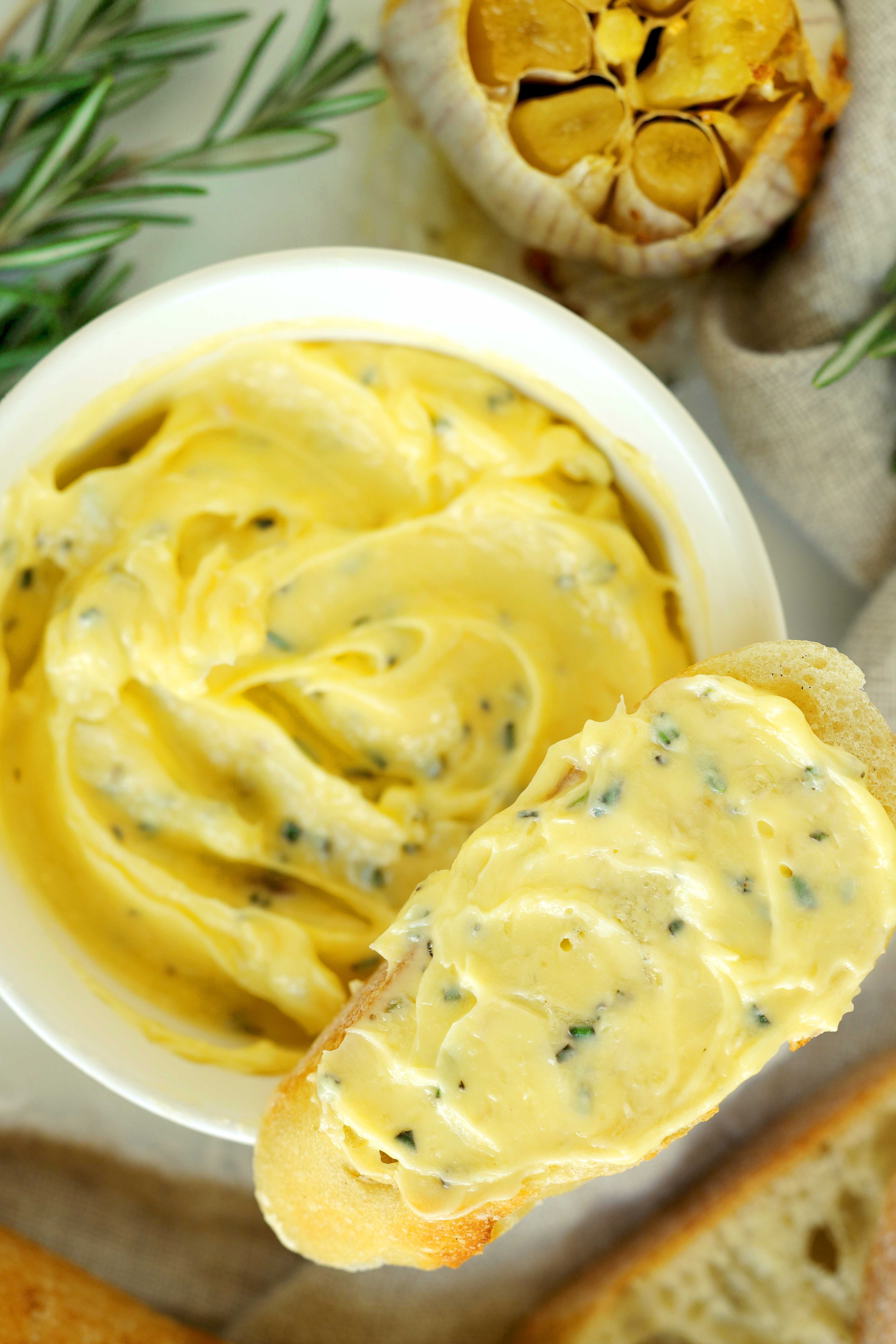 Close-up of a slice of crusty bread spread with creamy roasted garlic butter and herbs.