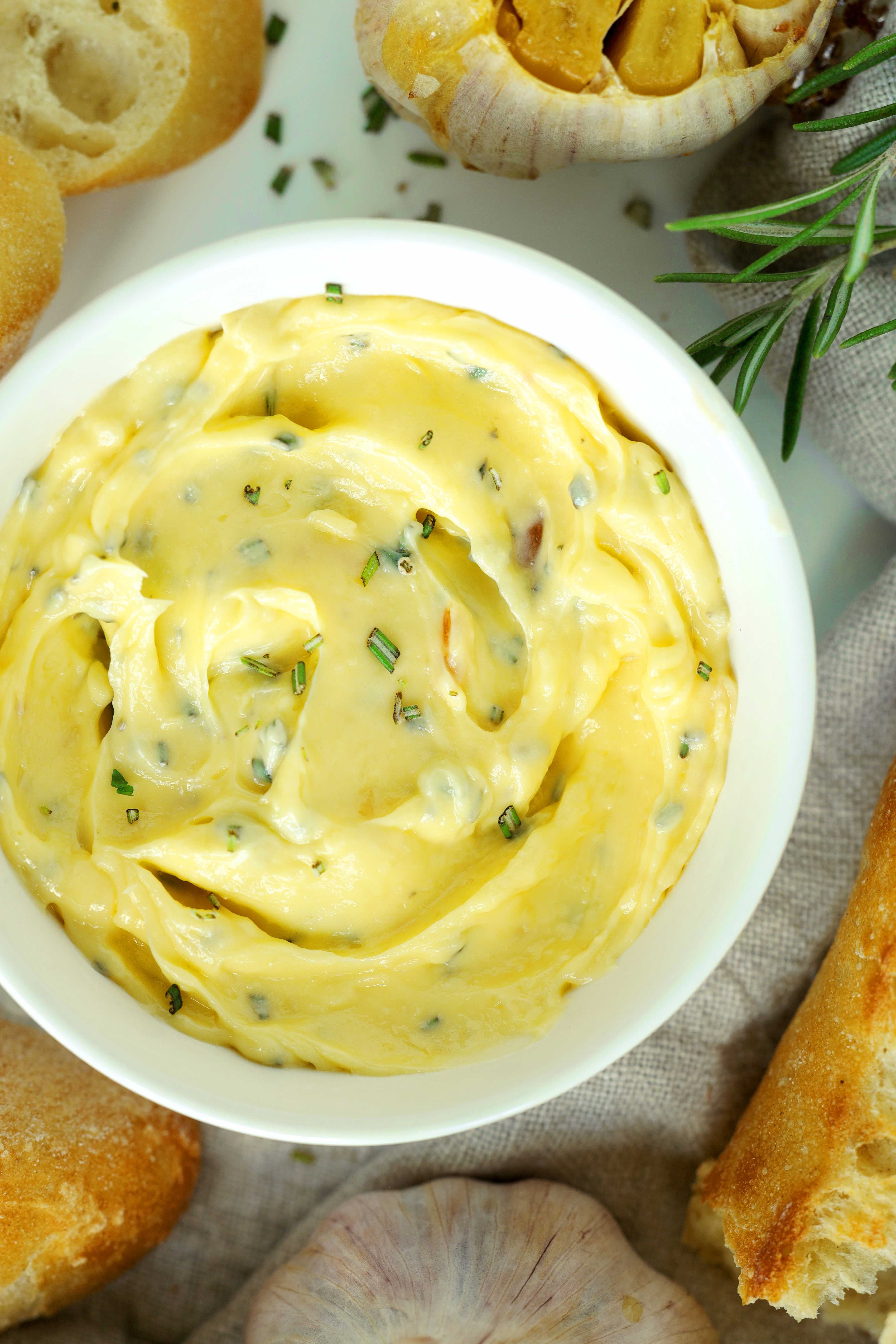 Bowl of homemade roasted garlic butter with chopped herbs, crusty baguette, and roasted garlic cloves.