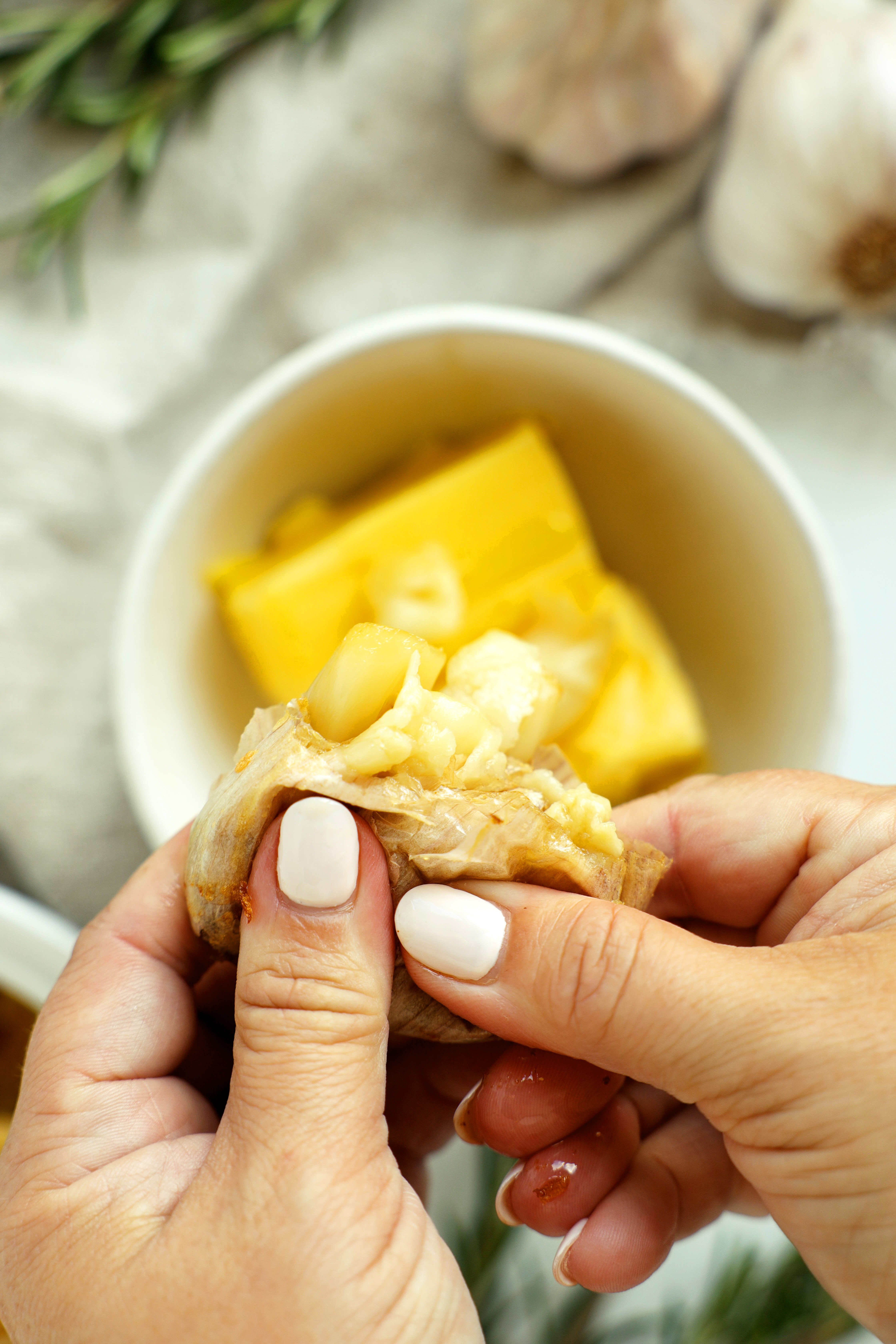 Hands squeezing tender roasted garlic cloves from their skins into a bowl of butter.
