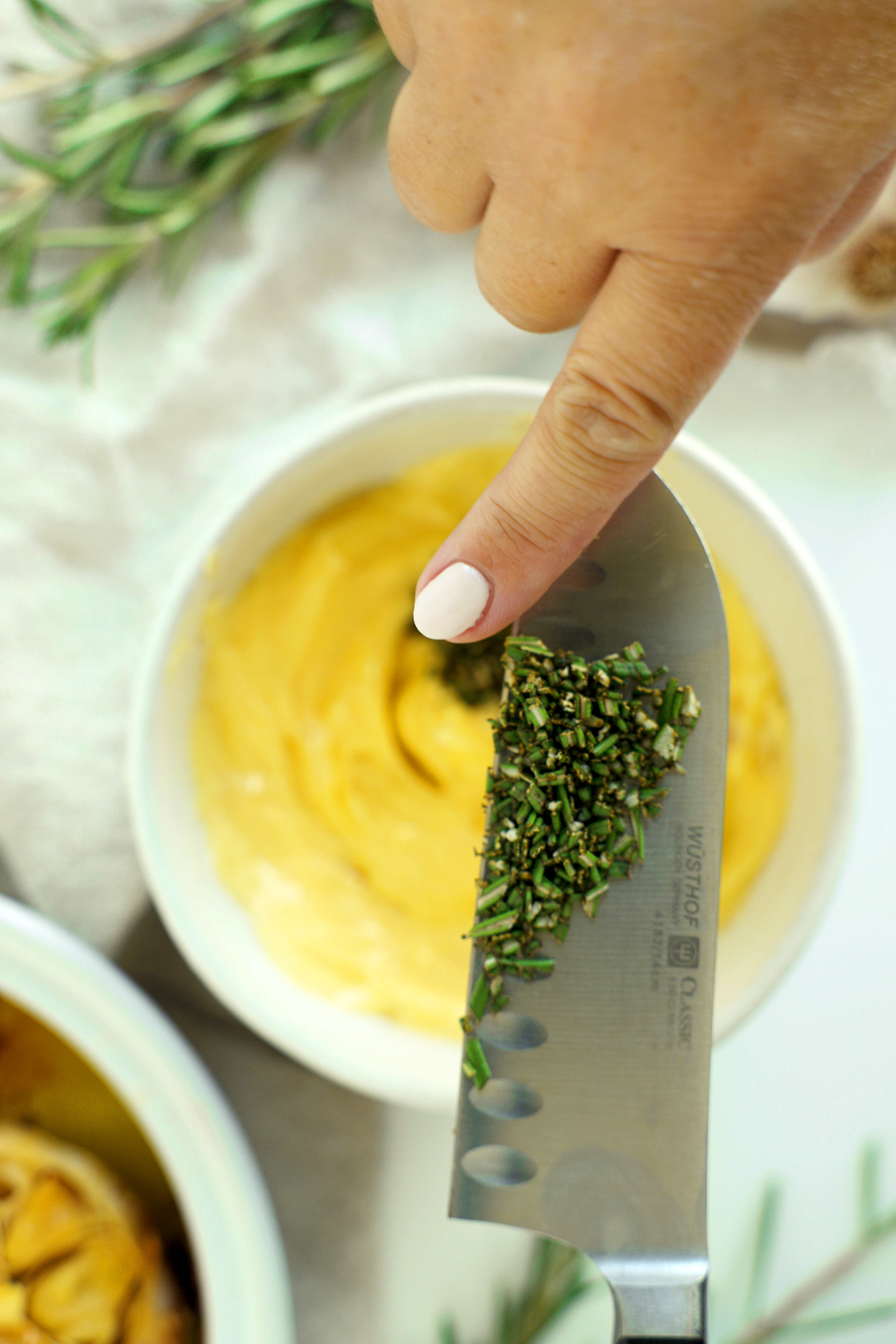 Chopped fresh rosemary on a knife blade being added to a bowl of garlic butter.