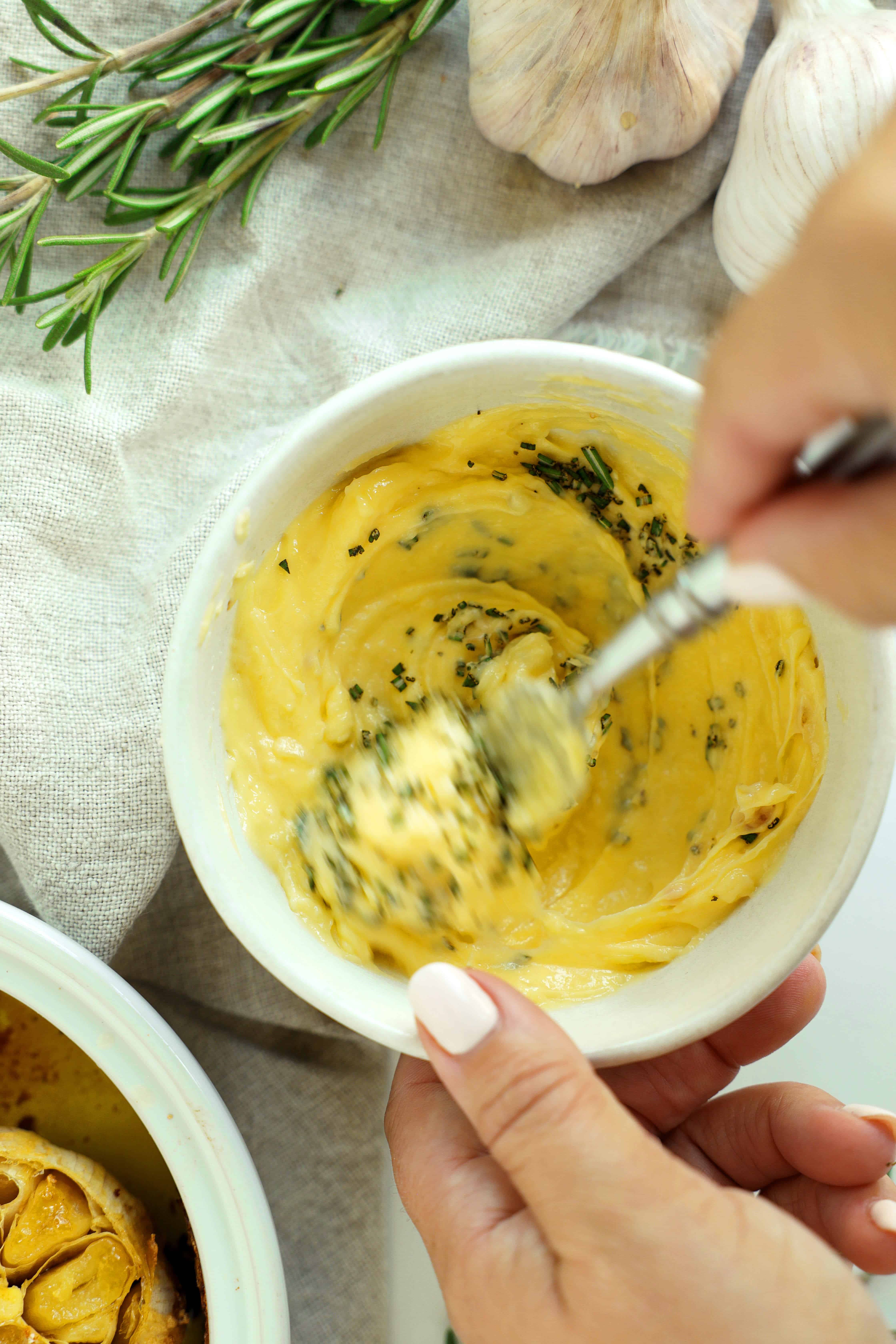 Hands stirring chopped rosemary into homemade roasted garlic butter with a fork.