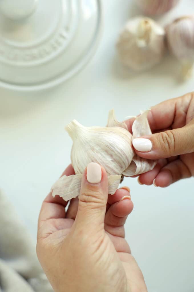 Hands peeling away the papery outer skin of a whole raw garlic bulb before roasting.