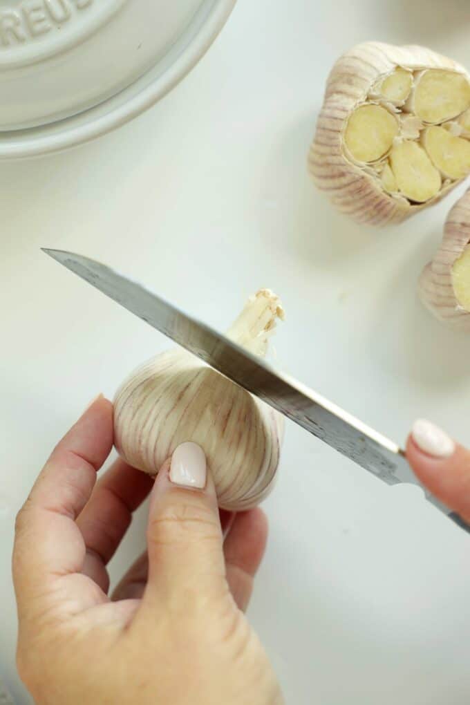 Knife slicing the top off a whole garlic bulb to expose the cloves for roasting.
