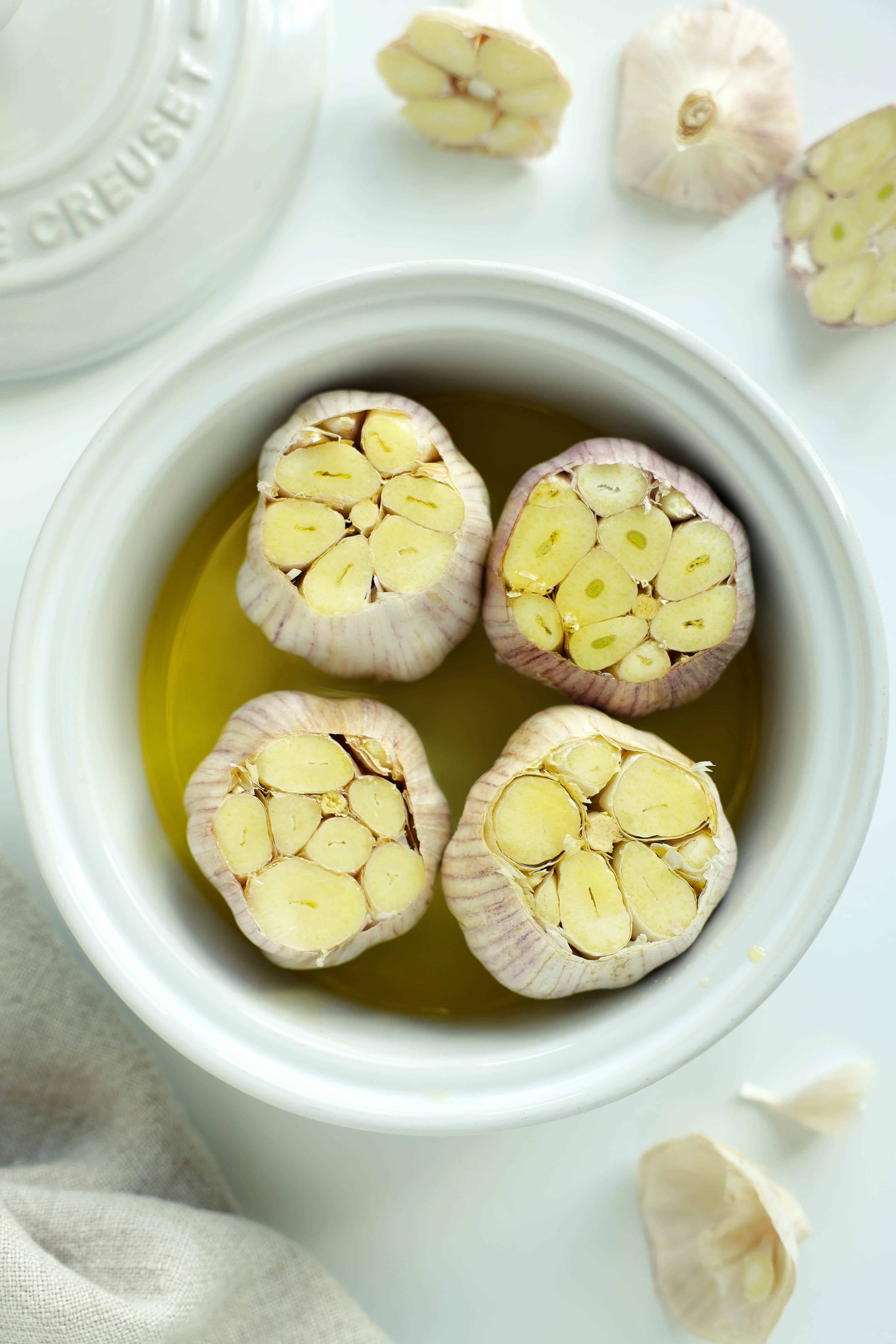 Four trimmed heads of raw garlic arranged cut-side up in olive oil in a white baking dish.